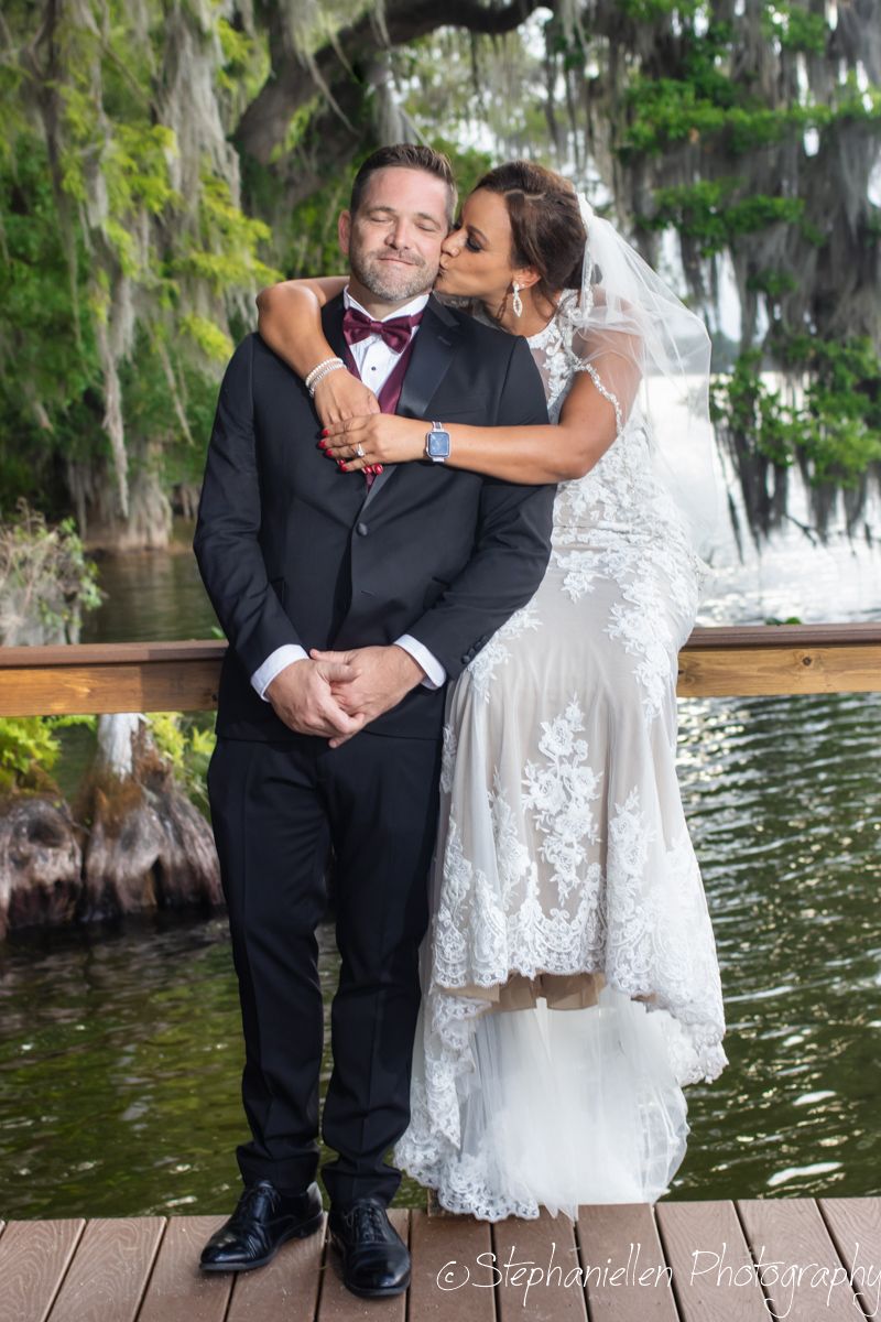 Bride in lace gown kissing groom on cheek on wooden dock near scenic waterfall.
