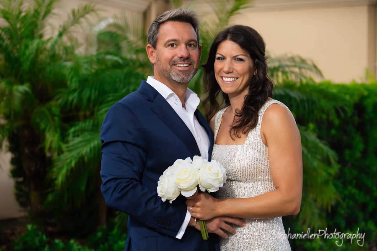 A happy couple poses outdoors among lush greenery, bride in a sequined dress holding white peony bouquet.