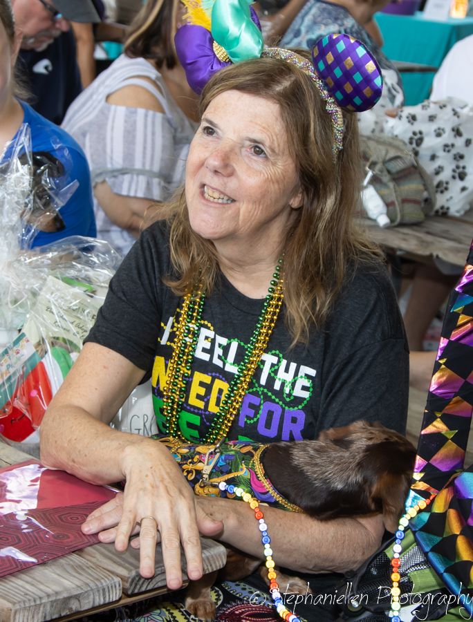 Woman wearing Mardi Gras beads and headband smiles while holding a small dachshund at a festive outdoor event.