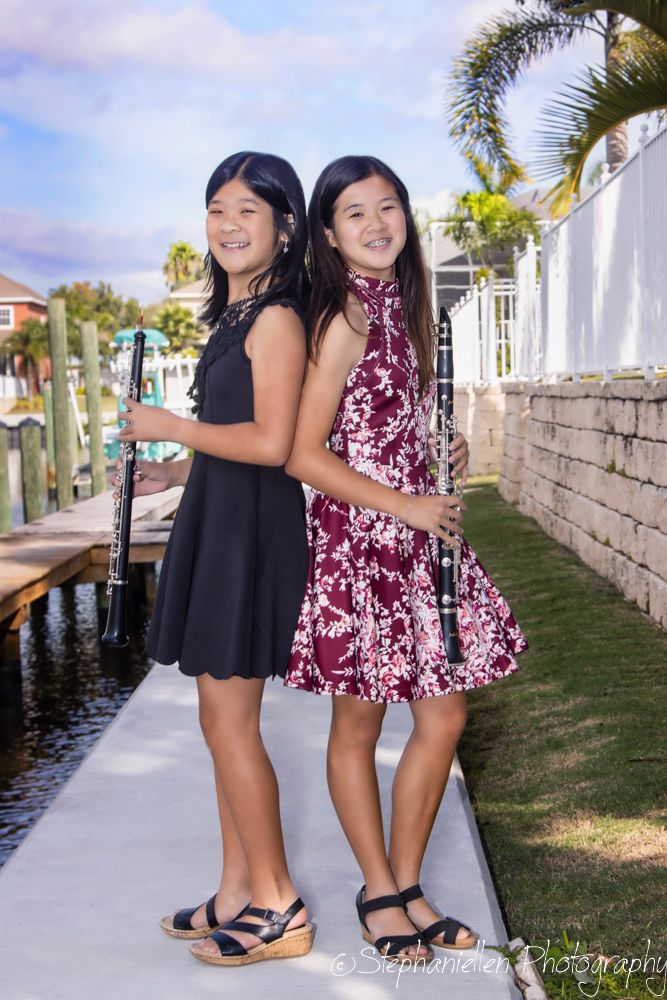 Two young girls in dresses holding clarinets pose back-to-back on a waterfront path on a sunny day.