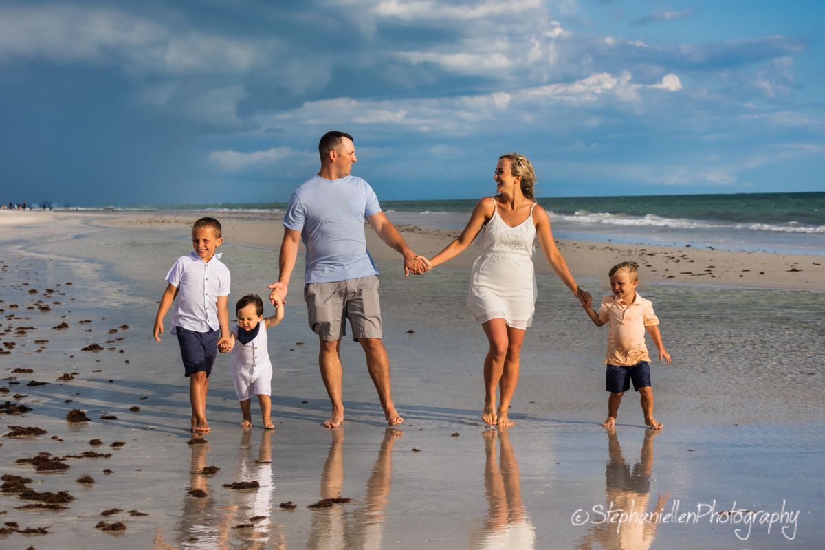 Family of five walking hand in hand on a sunny beach with dramatic storm clouds in the background.