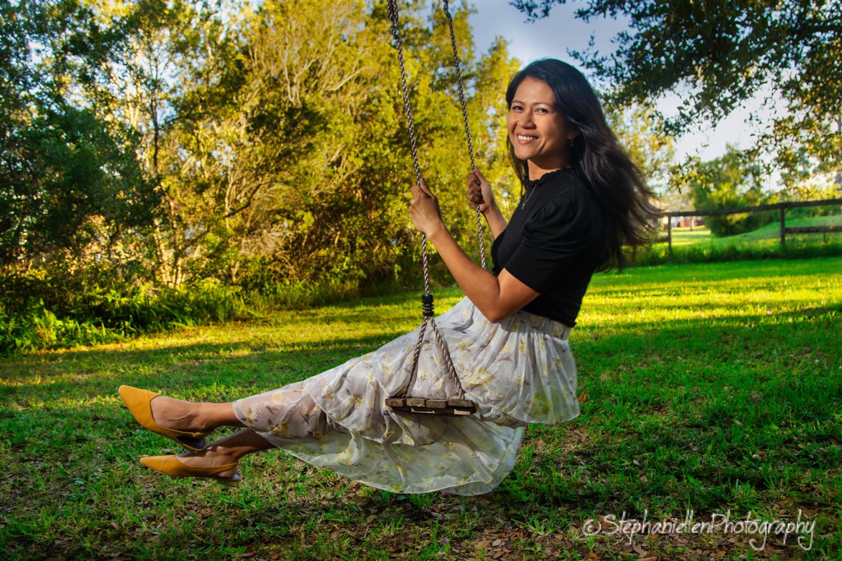 Young woman in black top and floral skirt sitting on a rope swing outdoors, smiling joyfully in a sunlit green park.
