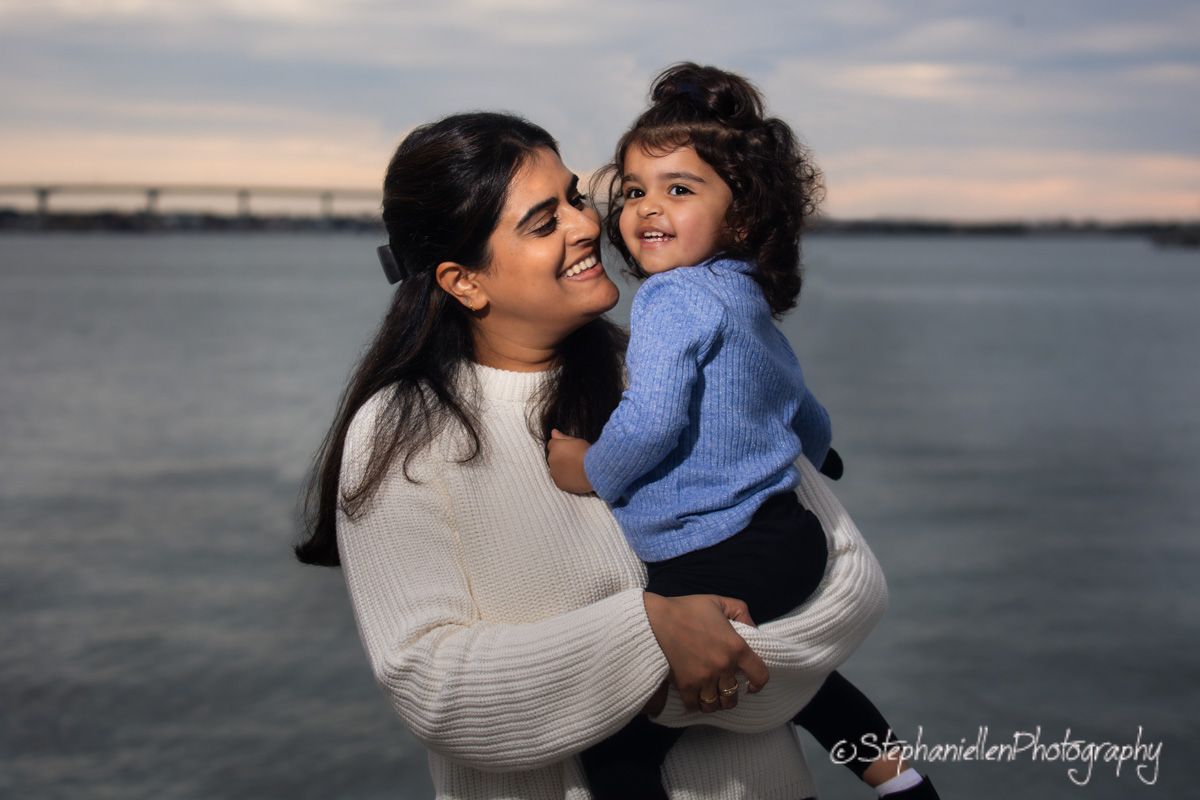 A woman in a white sweater lovingly holds a toddler in a blue shirt near a waterfront at dusk.
