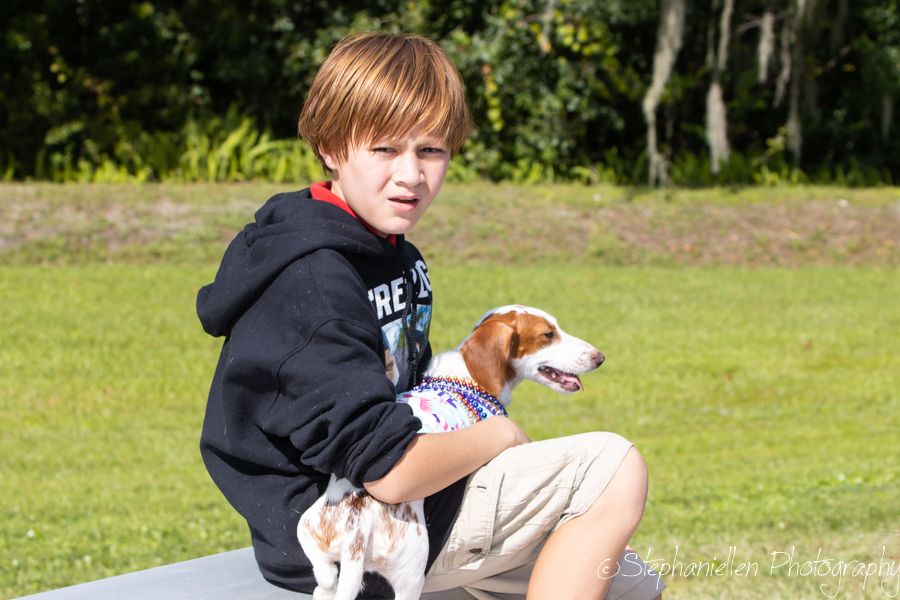 A young boy in a black hoodie sits outdoors holding a small beagle dog on his lap in a grassy park.
