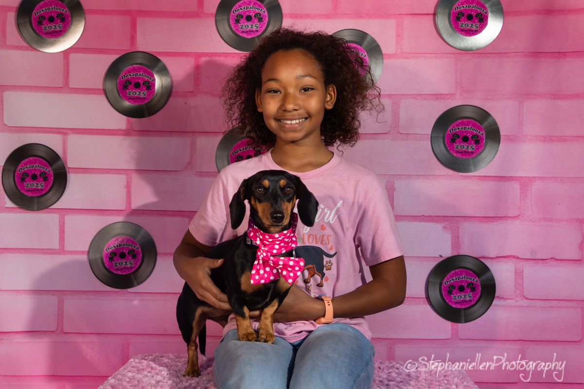 A smiling girl in a pink shirt holds a dachshund puppy with a polka dot bow in front of a pink brick backdrop.