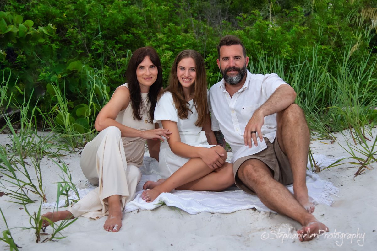 Family of three smiling together on white sand beach, surrounded by lush green vegetation in a professional photo.