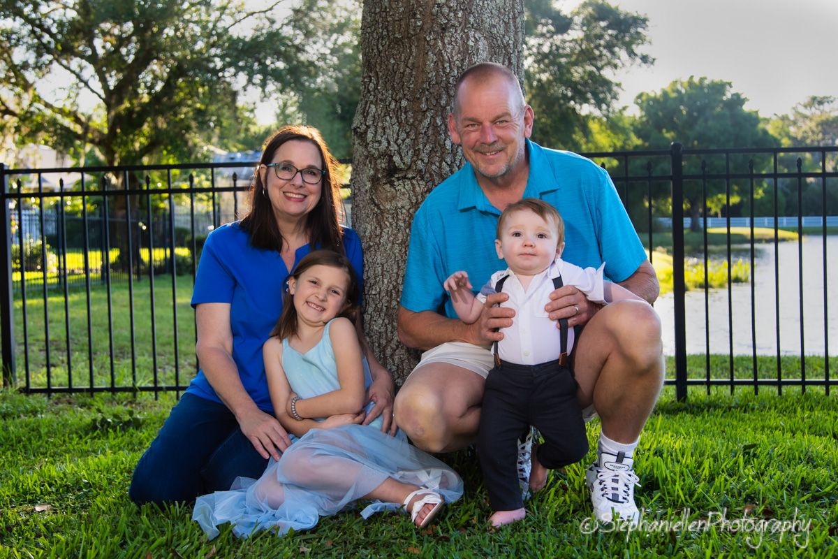 Family of four poses by a tree on green grass, parents with two children smiling outdoors.
