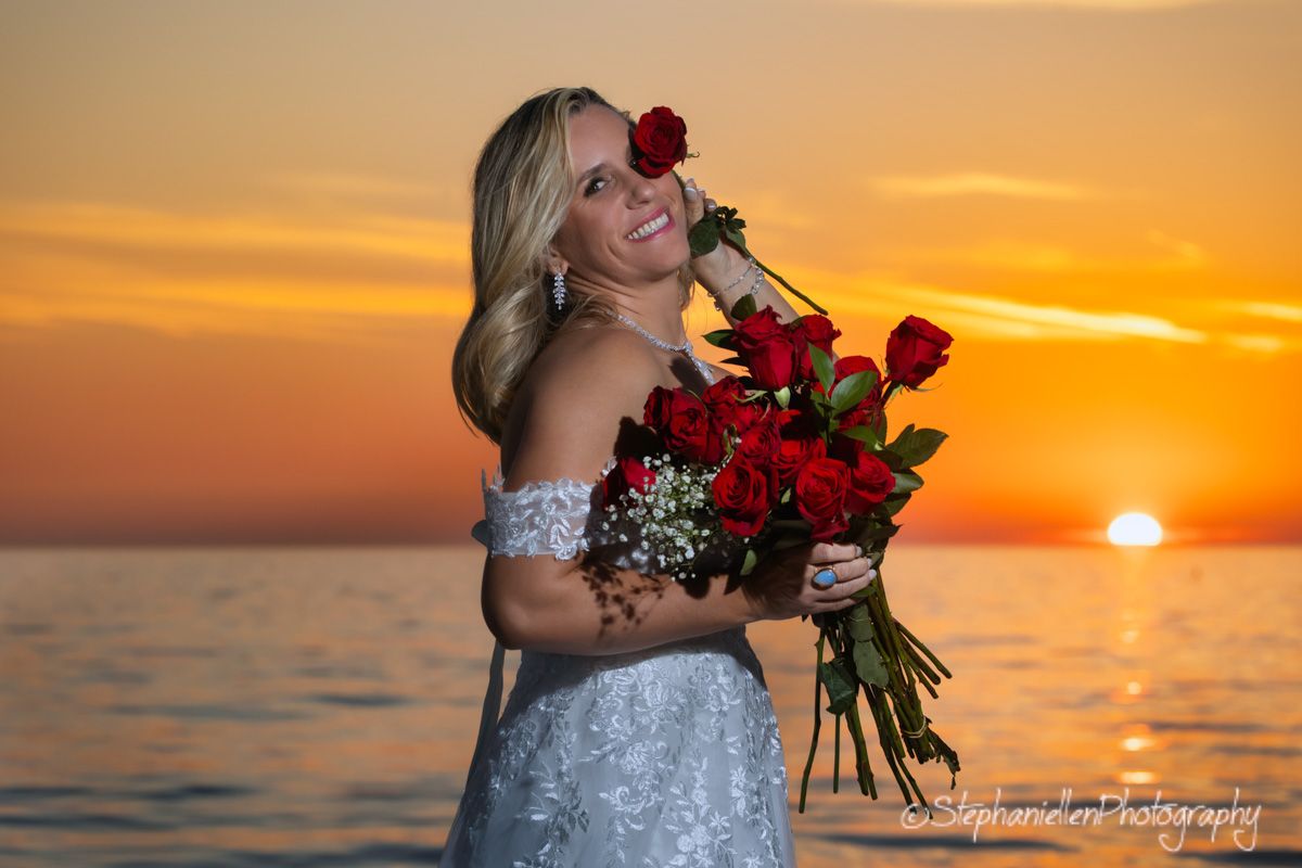 Smiling bride holding red roses bouquet at a beautiful ocean sunset.