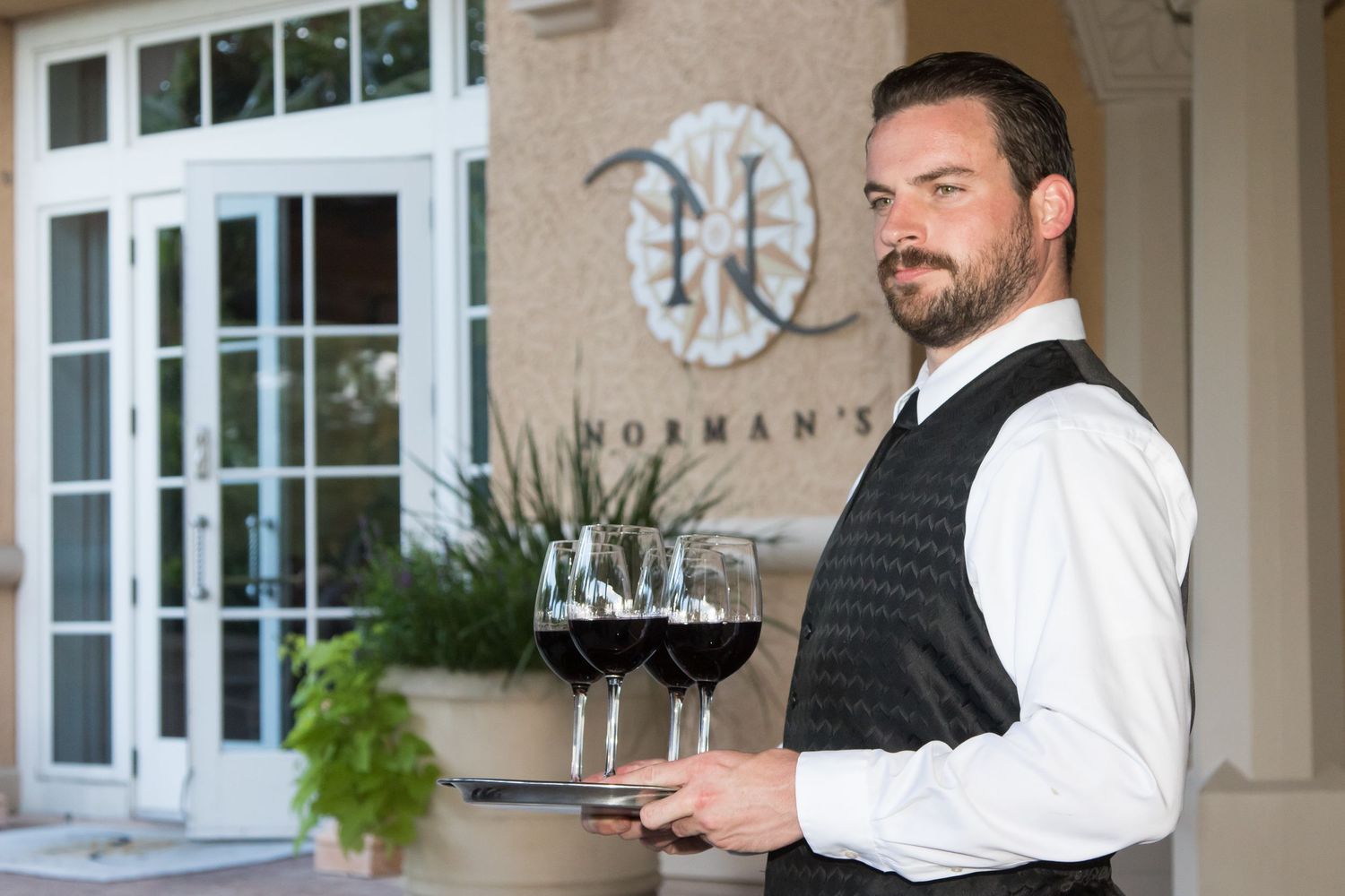 Bearded waiter in vest holds tray of red wine glasses outside Norman's restaurant.
