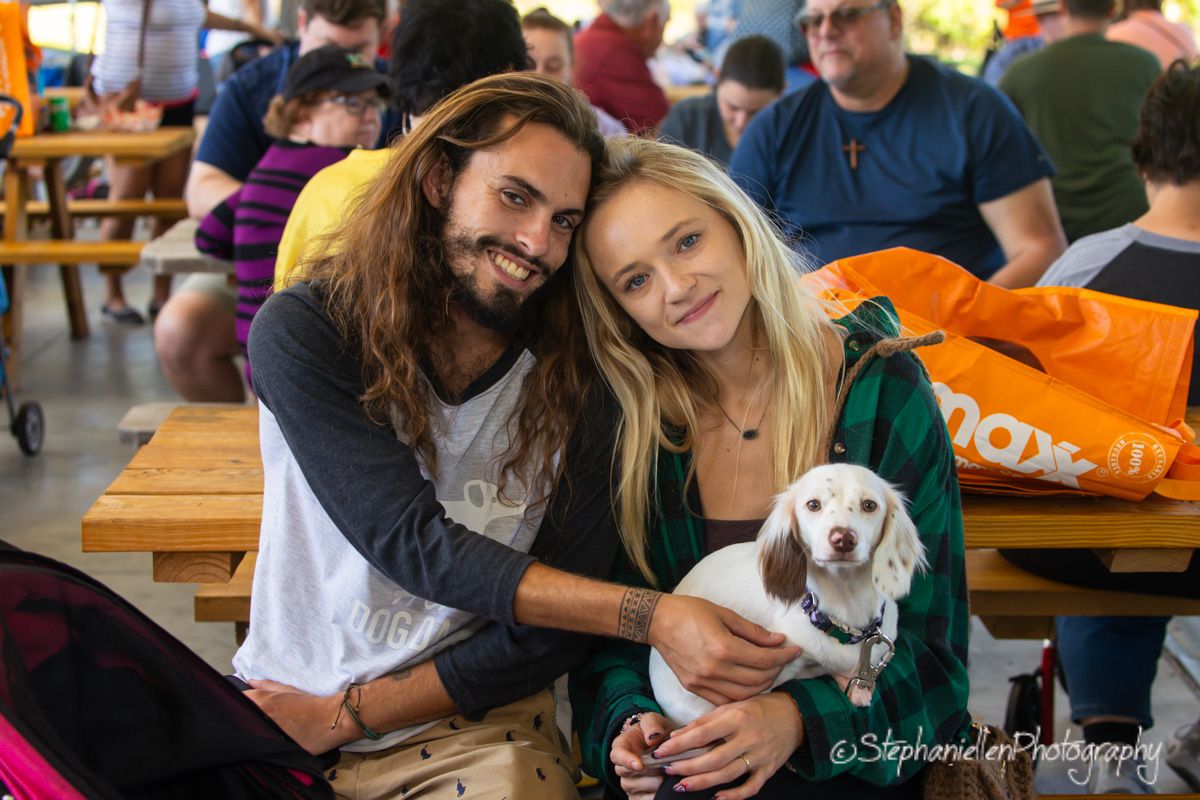 A couple smiles with their white dog at an outdoor gathering, the woman in green holding the pet affectionately.