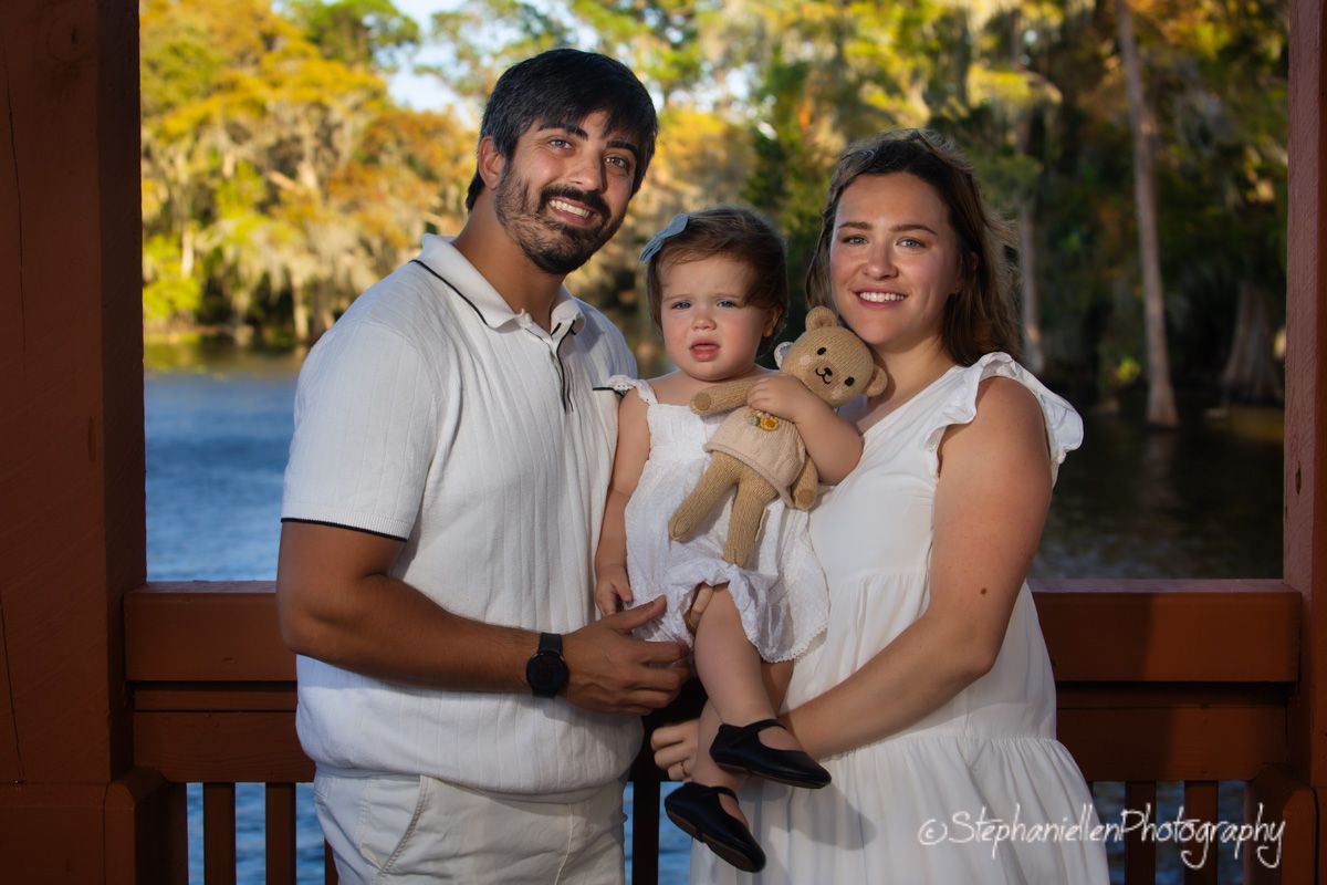 Happy couple in white outfits holding their baby and teddy bear near a scenic lakeside.