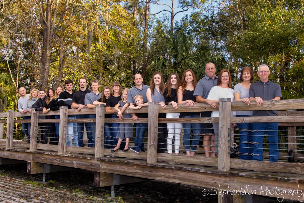 Large family group portrait on a wooden bridge surrounded by lush green trees outdoors.