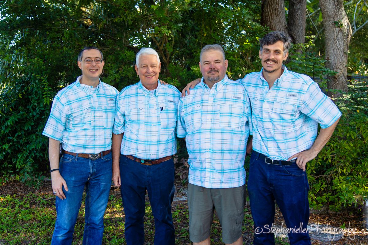 Four men in matching light blue plaid shirts smiling together outdoors among green trees in a family portrait.