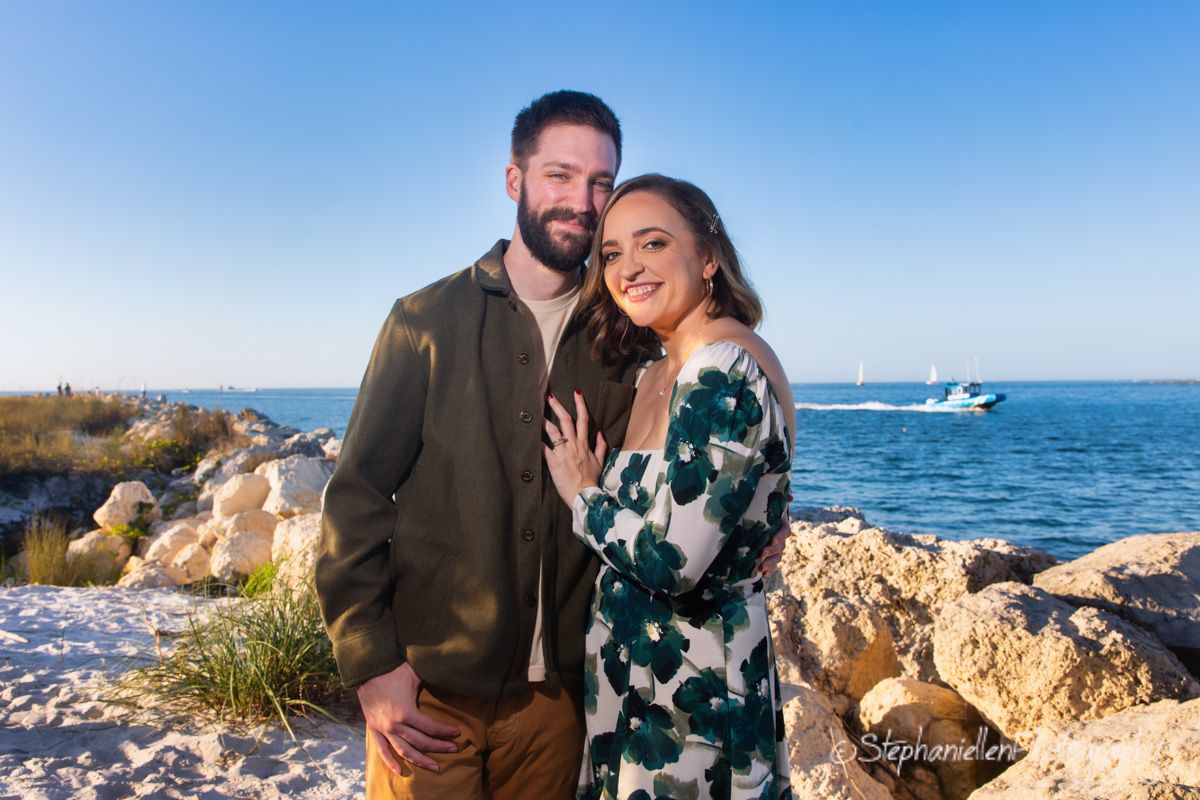 Couple smiling together on rocky shoreline with blue ocean and sailboat in background on sunny day.