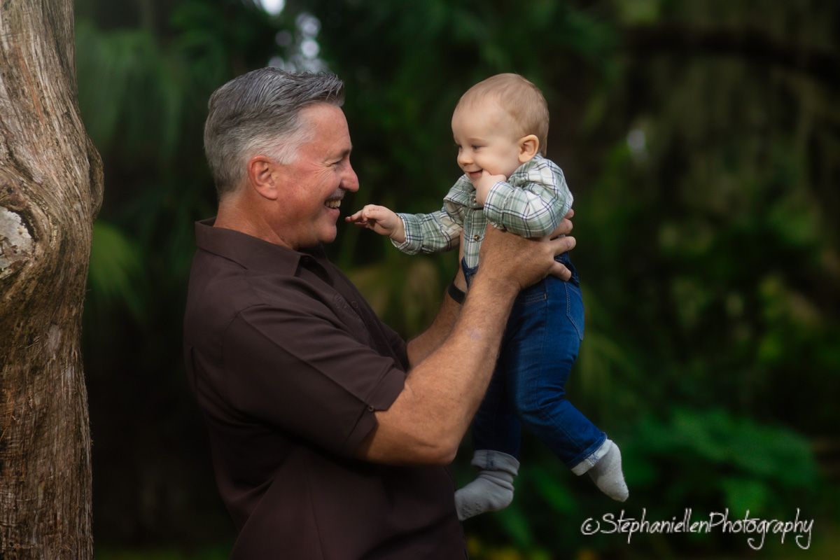 Man holding smiling baby up in lush green outdoor setting, captured by StephanieAnnPhotography.