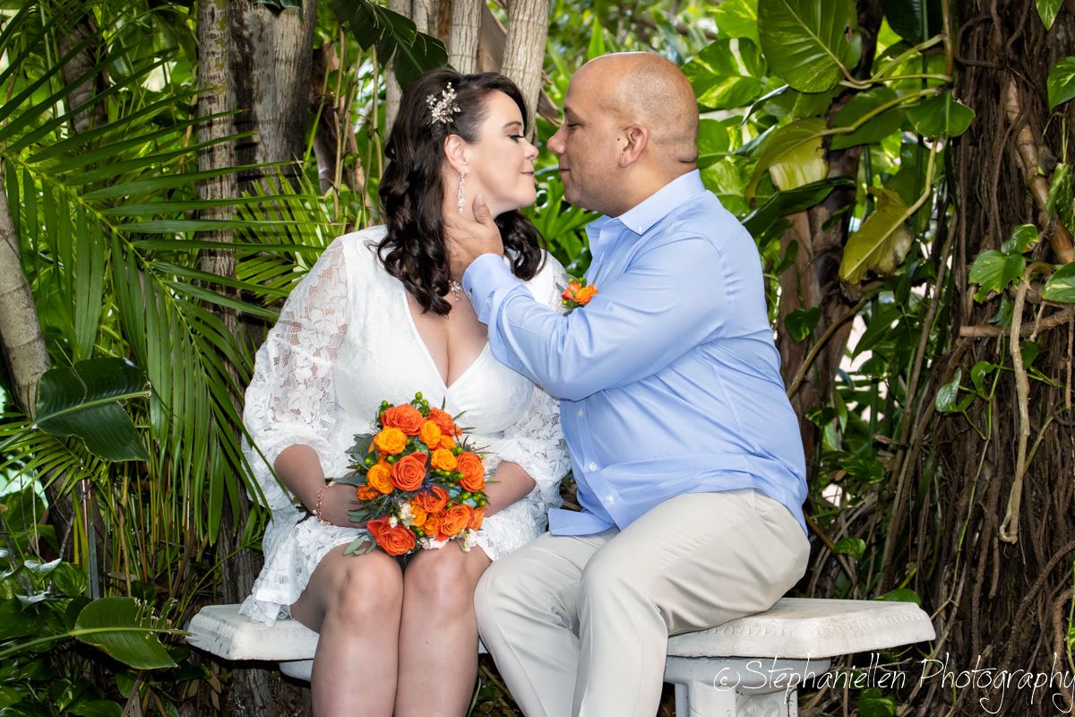 Bride and groom sharing an intimate moment on a bench surrounded by lush tropical greenery.