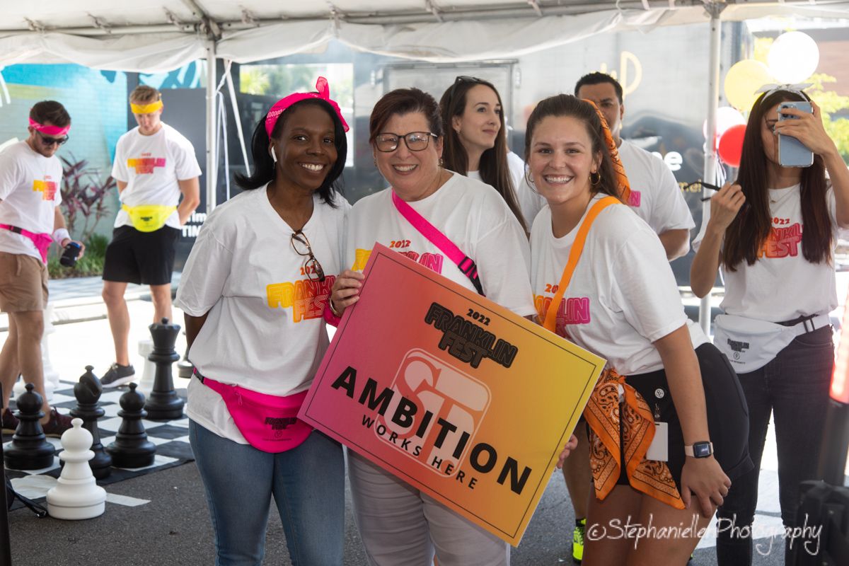 Three women smiling at an Ambition event holding a colorful sign outdoors.