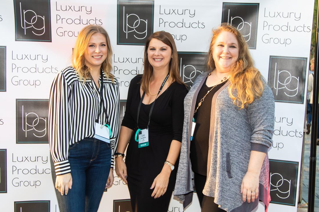 Three women smiling together at a Luxury Products Group event backdrop.