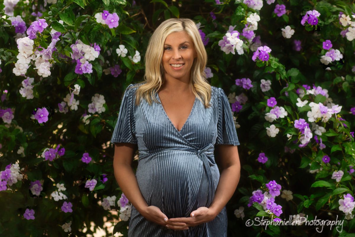 Smiling pregnant woman in blue dress standing before blooming purple and white flowers.