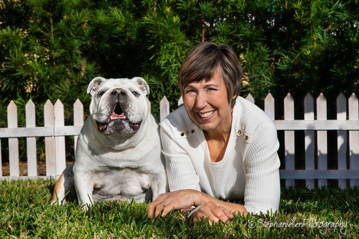 A woman smiles beside her English Bulldog on green grass in front of a white picket fence.