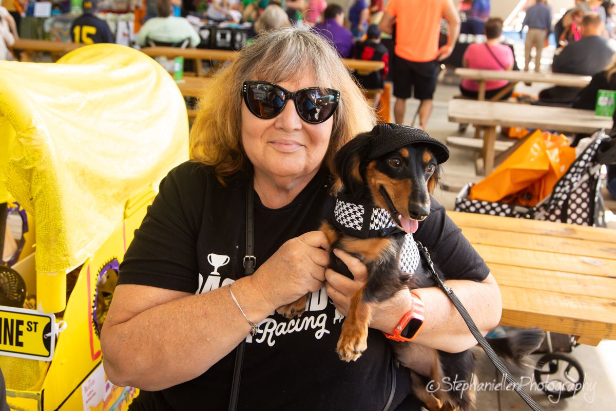 Woman in black sunglasses and t-shirt holds a dachshund wearing a bow tie at an outdoor dog event.