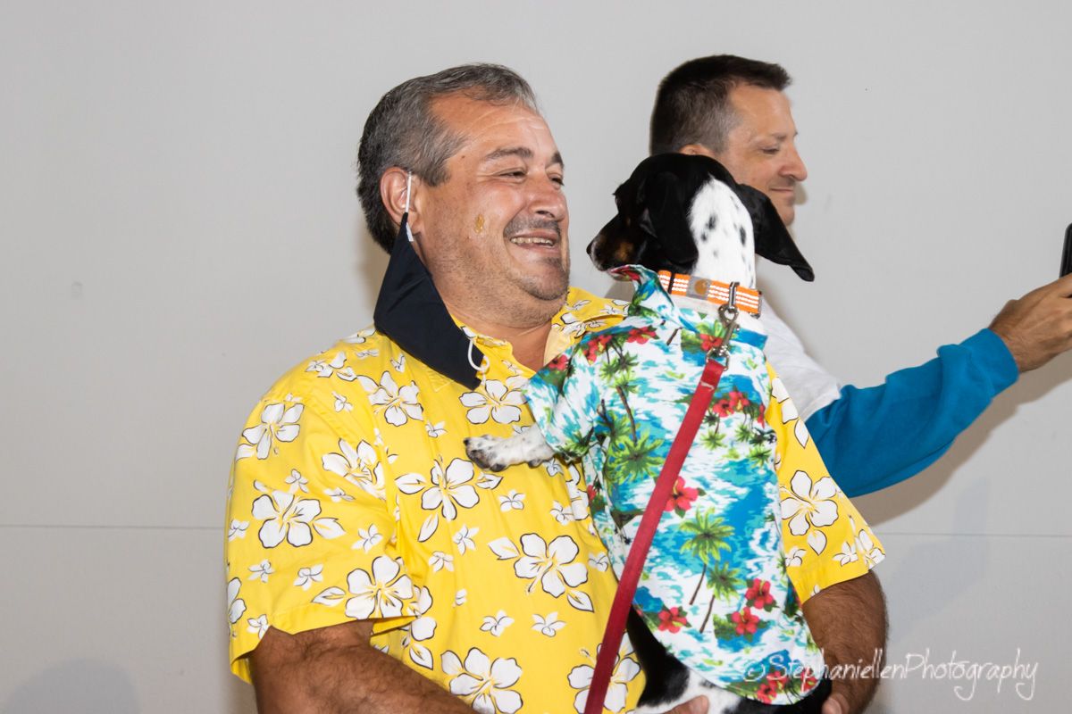 Man in yellow floral shirt laughing while holding a small dog dressed in matching Hawaiian shirt at a pet event.