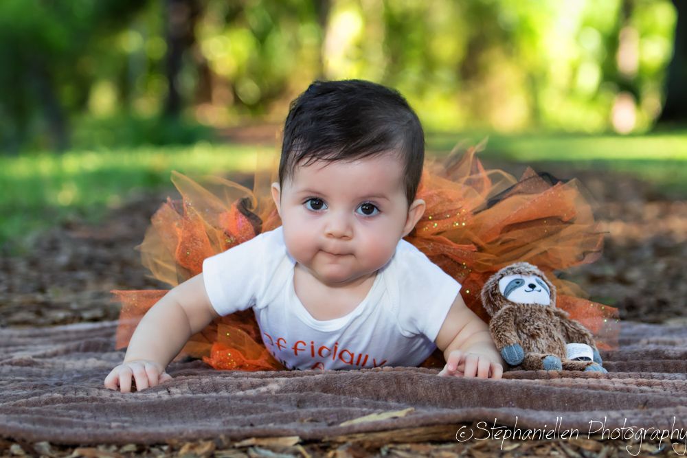 Adorable baby girl in orange tutu lying on blanket with stuffed sloth toy in park setting.