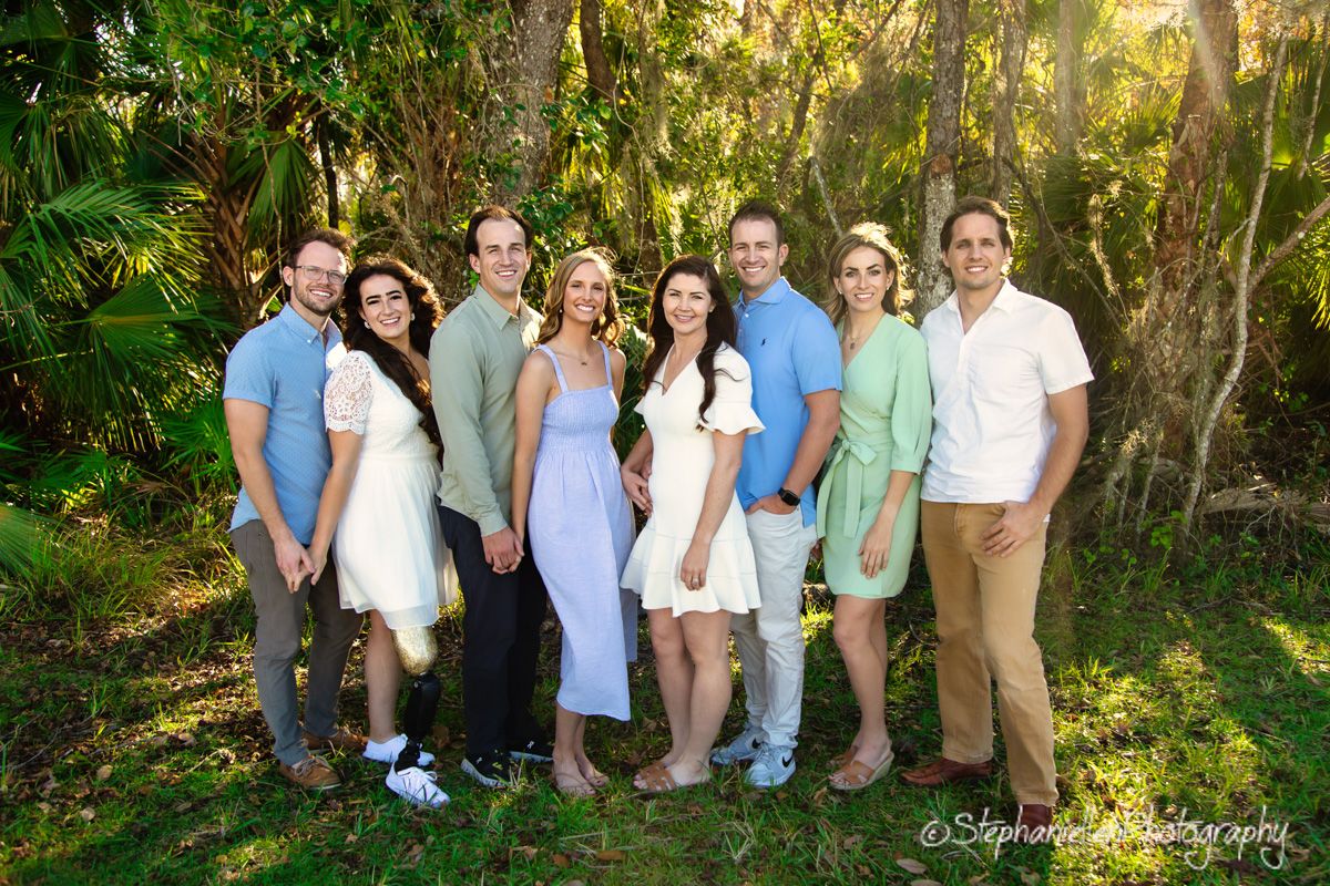 A happy family group of eight poses outdoors among lush tropical greenery in coordinated pastel outfits.
