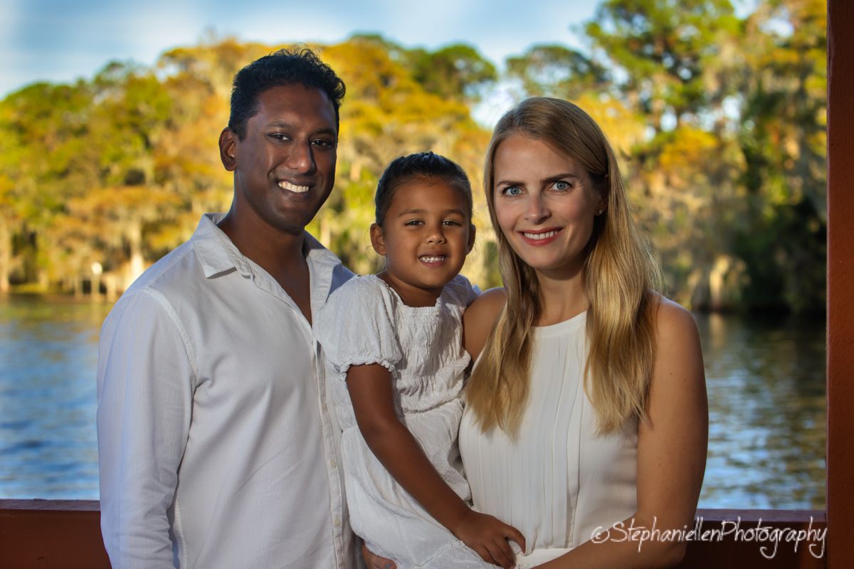 Smiling interracial family of three posing outdoors near a lake, wearing white outfits.
