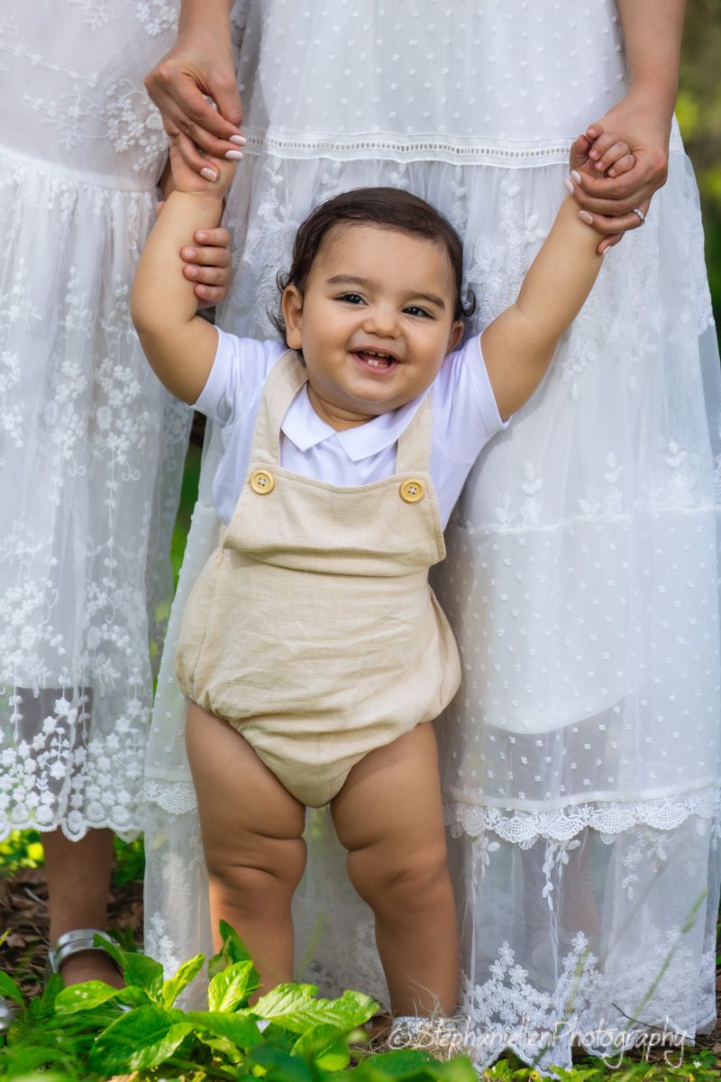 A happy baby in tan overalls stands supported by hands in a sunny outdoor garden setting.