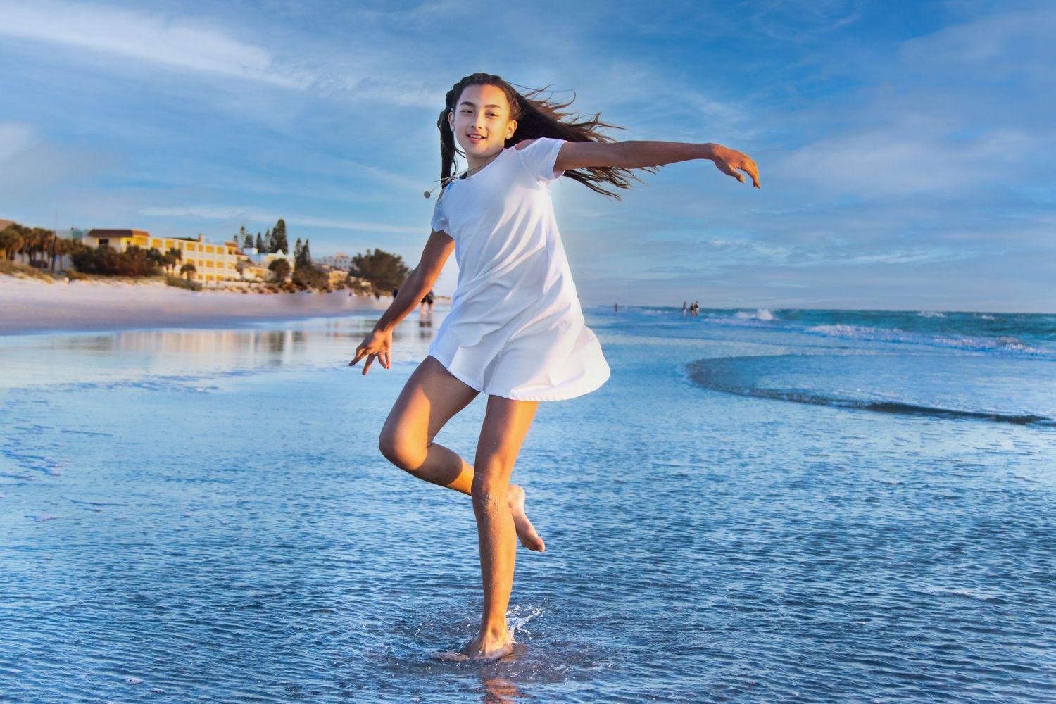 A joyful girl in a white dress dances barefoot on a sunny beach with gentle waves and blue sky.