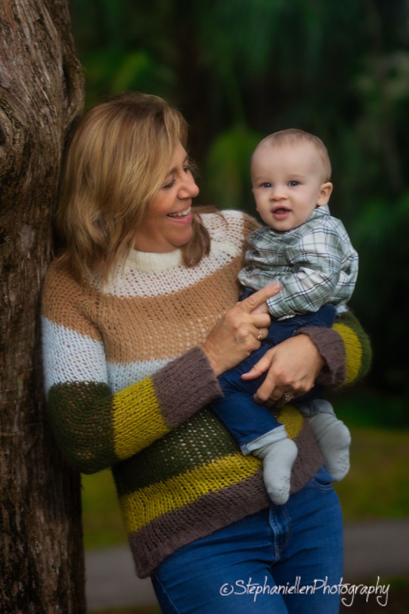 A smiling woman in a striped sweater holds a happy baby boy outdoors near a tree in a lush green park.