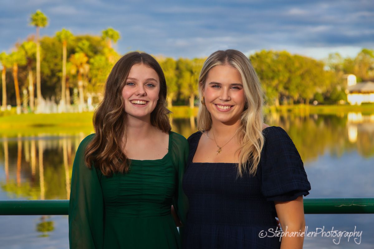 Two smiling young women in green and navy dresses pose outdoors near a scenic lake with palm trees at sunset.