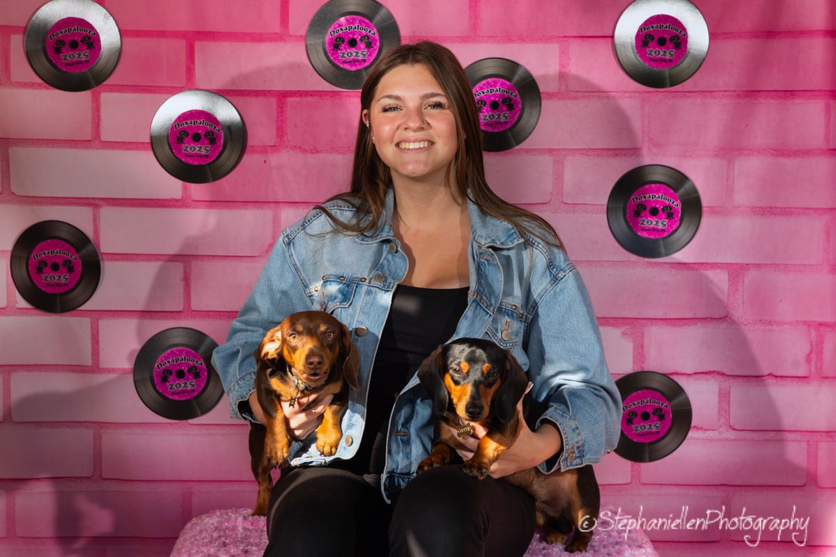 Woman in denim jacket smiling while holding two dachshund dogs in front of a pink brick wall with vinyl record decorations.