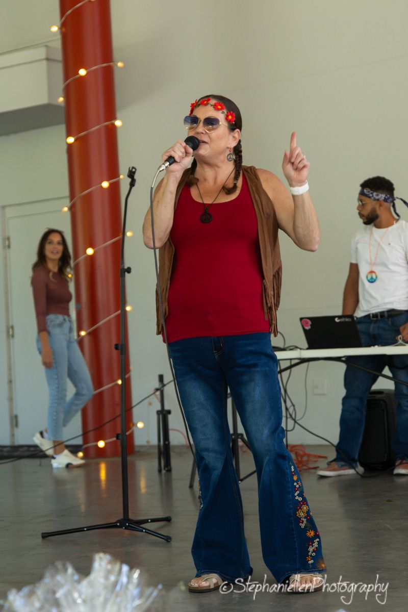 A woman in retro 70s outfit with flared jeans sings into microphone at outdoor festival event.