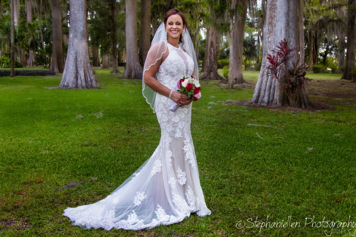 Bride in fitted lace mermaid gown with veil holds red and white bouquet standing in a palm tree-lined lawn.