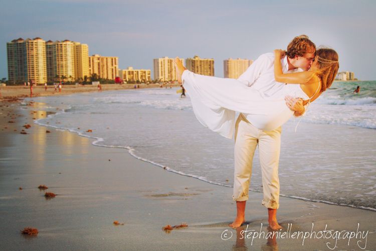 Groom lifting bride in white dress on a sunny beach with city skyline behind them.