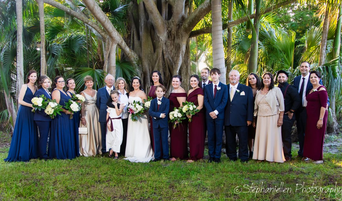 Large wedding party poses outdoors under tropical trees, bridesmaids in navy and burgundy gowns with white bridal couple.