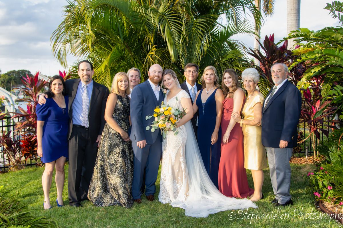 Wedding party poses outdoors with tropical greenery, bride holding yellow bouquet in white gown surrounded by family.
