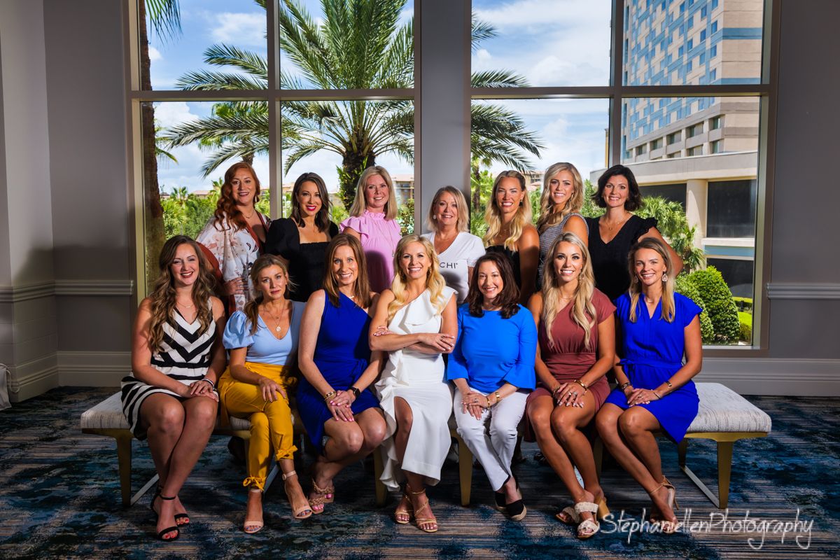 Group of fourteen women pose together in hotel lobby with palm trees in background.