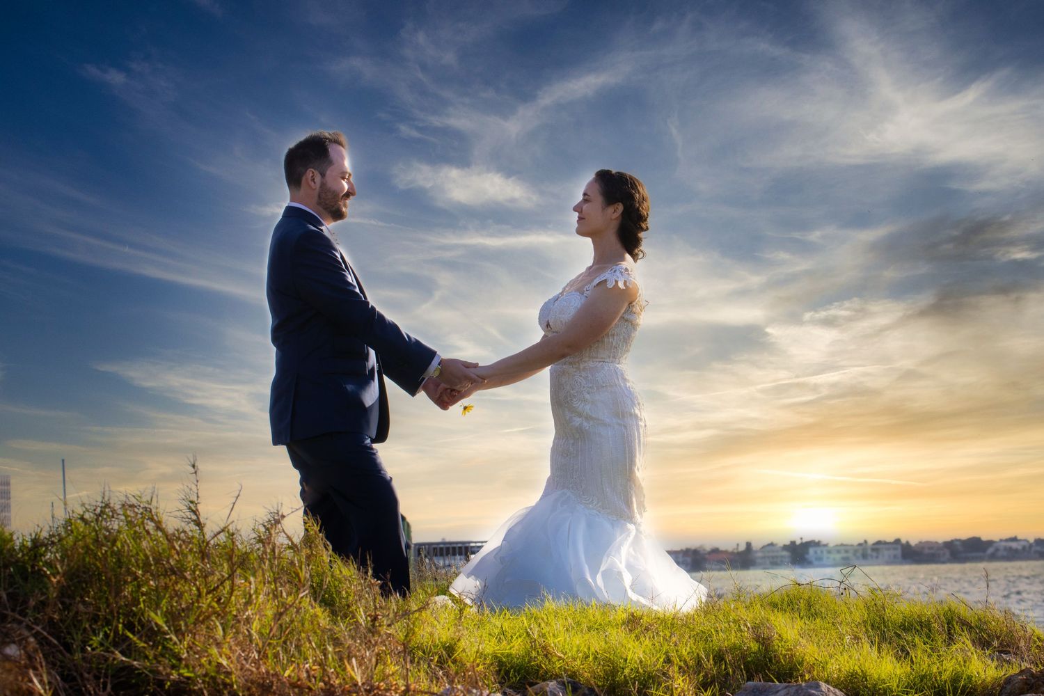 Bride and groom hold hands smiling at sunset beside waterfront on grassy hill.