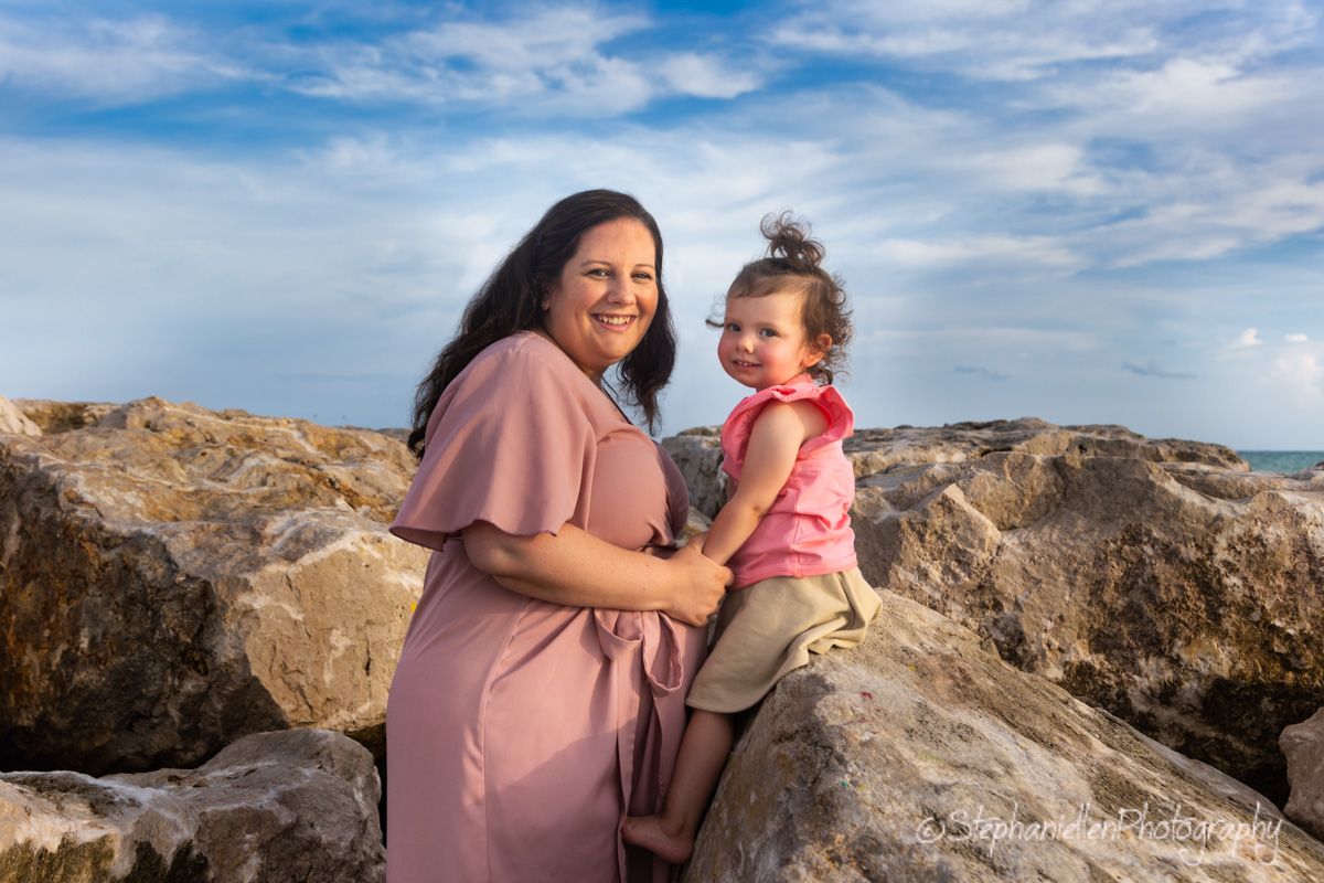 Smiling mother in pink dress holding young child on rocky beach with blue sky backdrop.