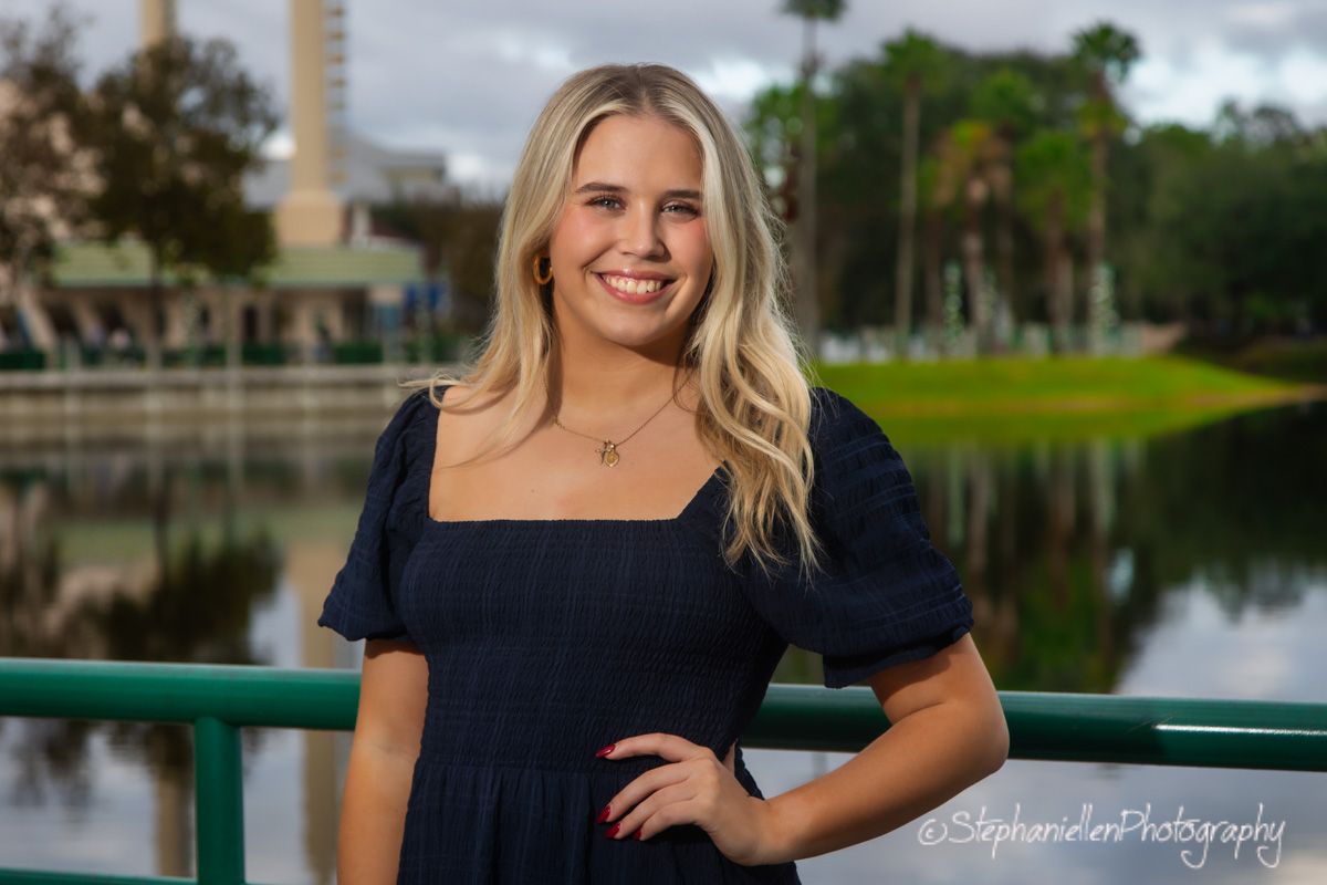 Young blonde woman smiling in navy dress poses by a scenic pond with lush greenery and buildings in background.