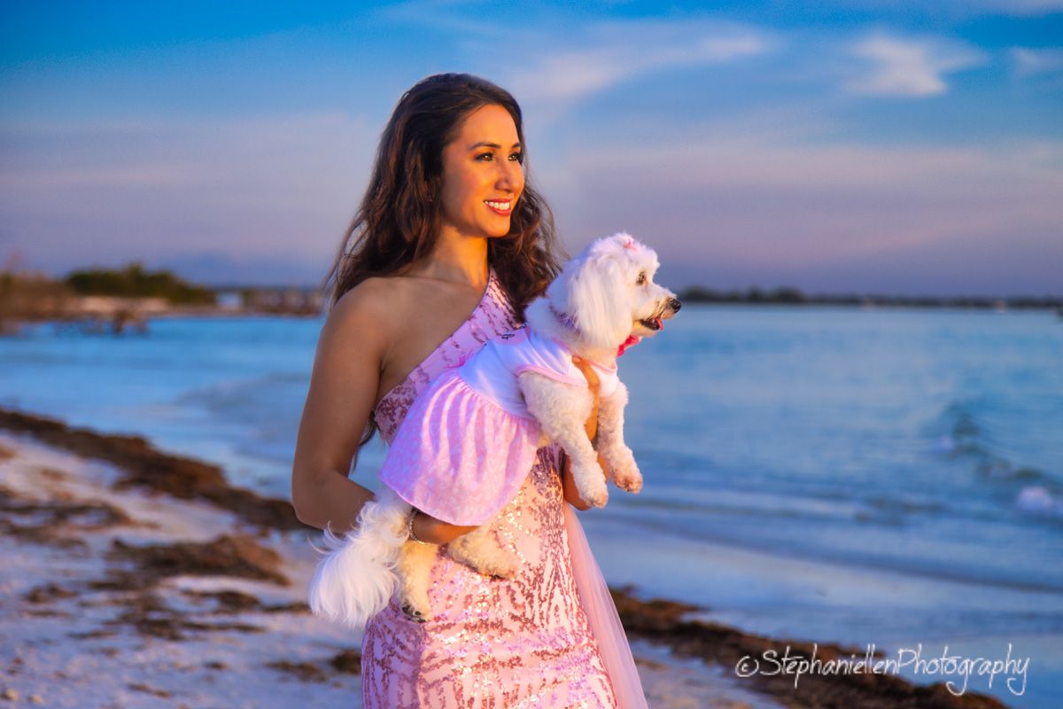 Smiling woman in pink dress holds white fluffy dog on beach at sunset.