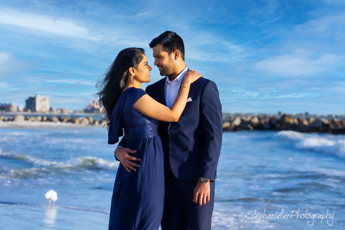 Elegant couple in coordinating blue outfits embracing on a sunny beach for an engagement photoshoot.