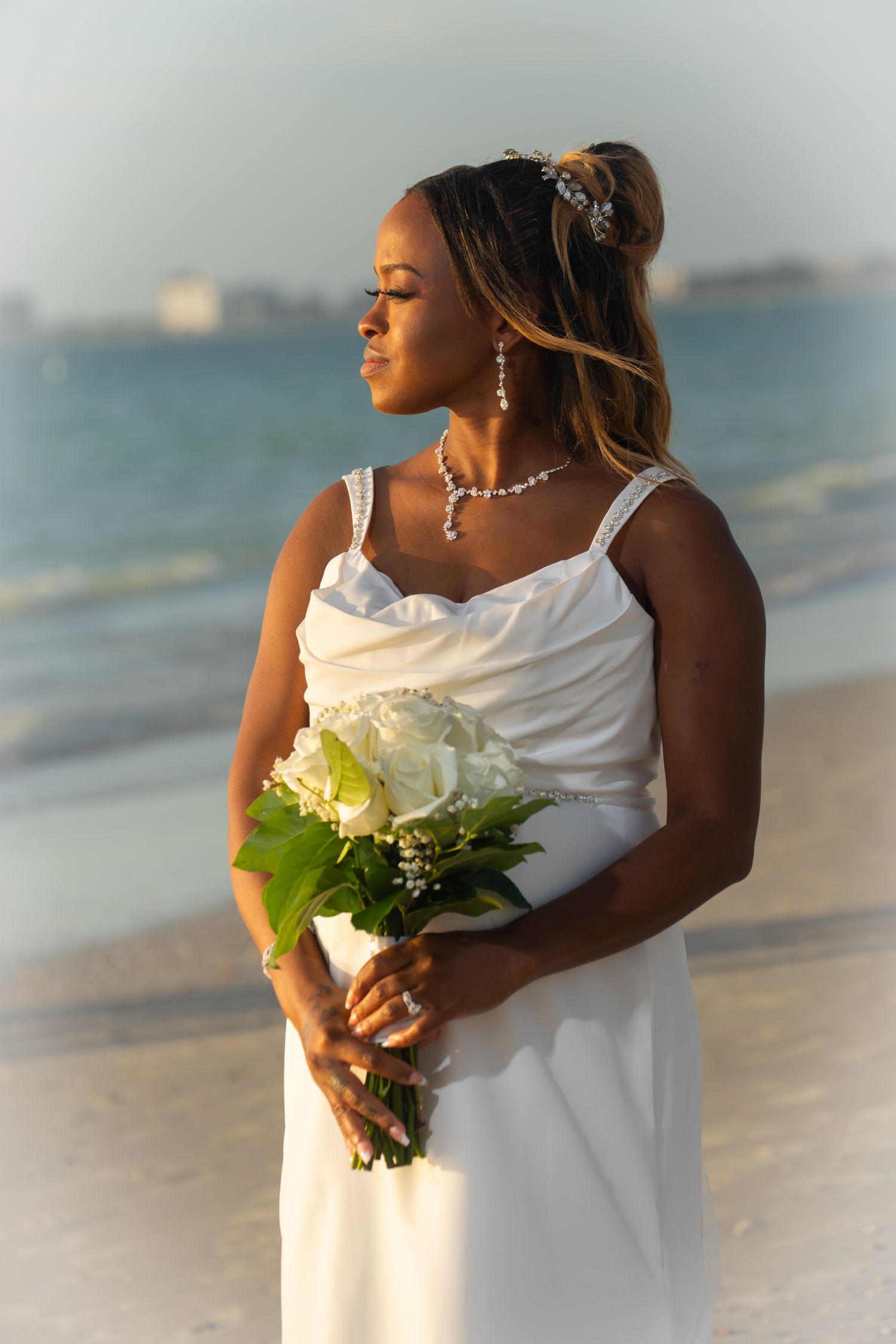 Elegant bride in white dress holding white rose bouquet on sunny beach.