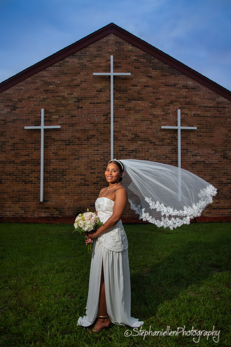 Bride in white gown with flowing veil poses before a brick church with crosses.