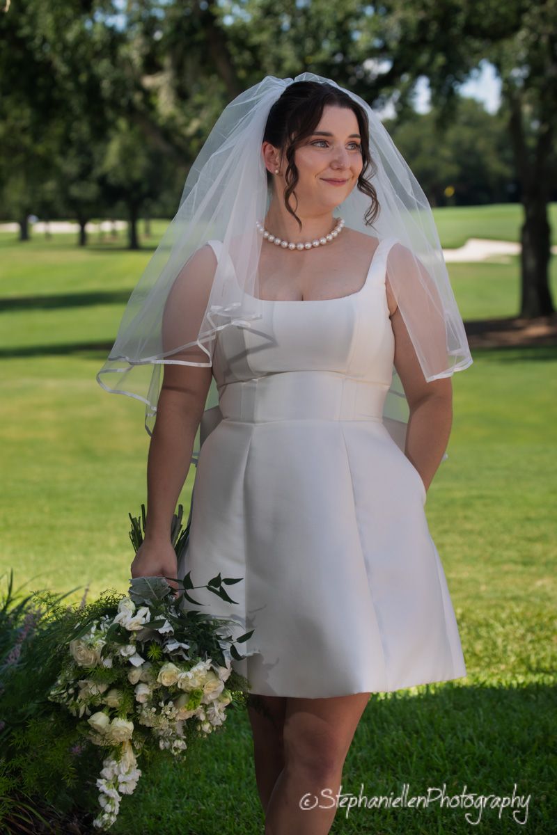 Bride in short white wedding dress with veil holding white floral bouquet on golf course.
