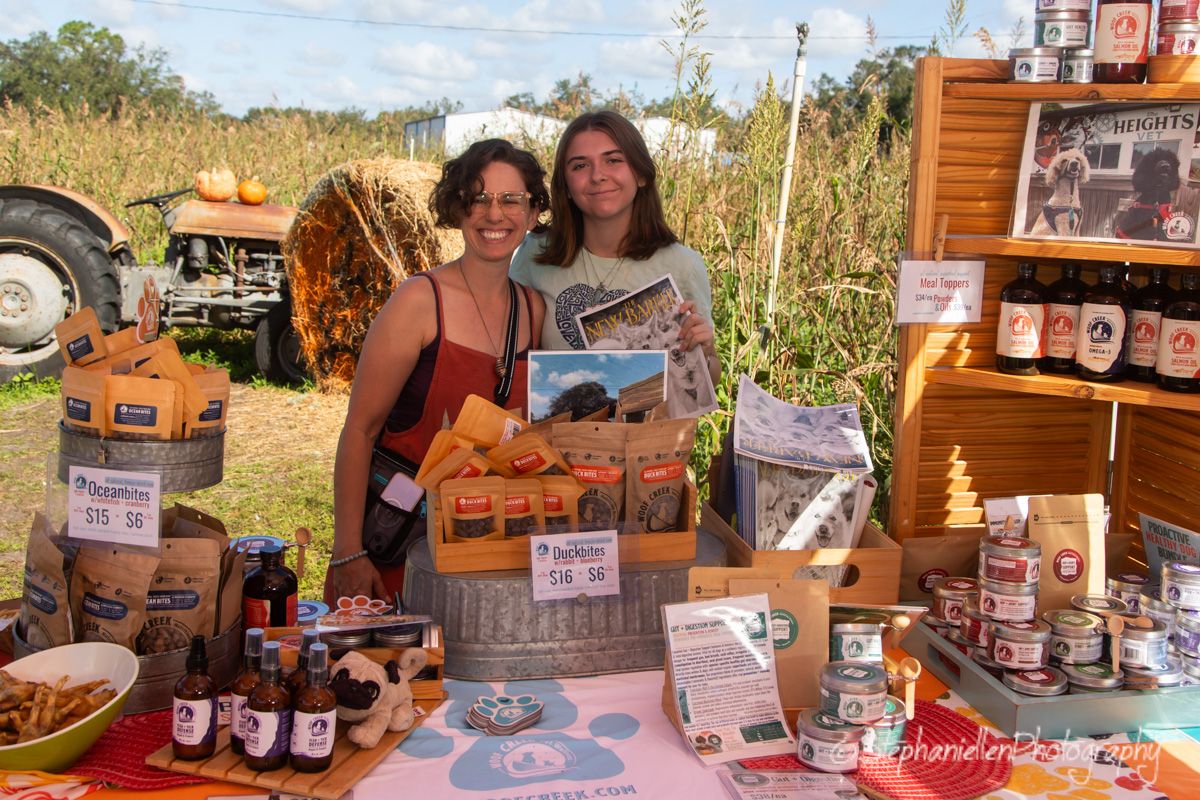 Two women smiling at a rustic farm market booth with jams, sauces, and country goods for sale.