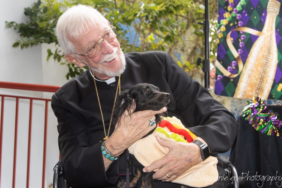 Elderly man in black clergy attire smiling while holding a small dog dressed in a hot dog costume at an event.